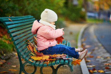 Adorable toddler girl sitting on the bench and playing with fallen leaves