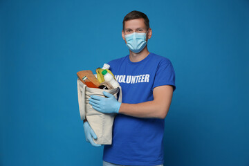 Male volunteer in protective mask and gloves with products on blue background. Aid during...