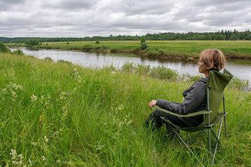 Young woman sitting in the outdoor chair on the bank of Ugra river. Ugra national park, Kaluga oblast, Russia.