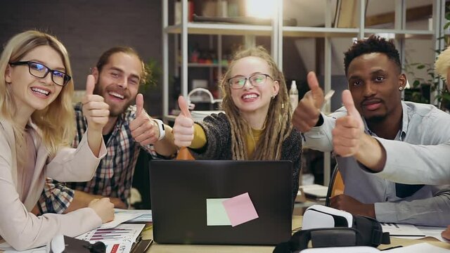 Likable positive creative lucky young multiethnic office colleagues showing thumbs up and looking at camera in modern meeting room