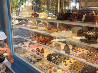 Small baby attached to his mother is before the pastry shop and checking the deserts in the window as if he can eat. Baby picks up a desert from the pastry shop.