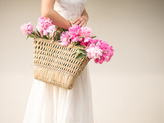 Boho girl holding pink peonies in straw basket. Stylish hipster woman in bohemian floral dress gathering peony flowers on white backgrounds. International Womens Day. Wedding decor and arrangement