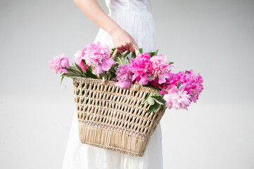 Boho girl holding pink peonies in straw basket. Stylish hipster woman in bohemian floral dress gathering peony flowers on white backgrounds. International Womens Day. Wedding decor and arrangement