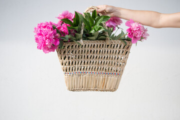 Boho girl holding pink peonies in straw basket. Stylish hipster woman in bohemian floral dress gathering peony flowers on white backgrounds. International Womens Day. Wedding decor and arrangement