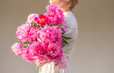 Boho girl holding pink peonies in straw basket. Stylish hipster woman in bohemian floral dress gathering peony flowers on white backgrounds. International Womens Day. Wedding decor and arrangement