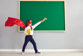 Back to school. Schoolgirl in a superhero costume on the background of a green school board in the classroom.