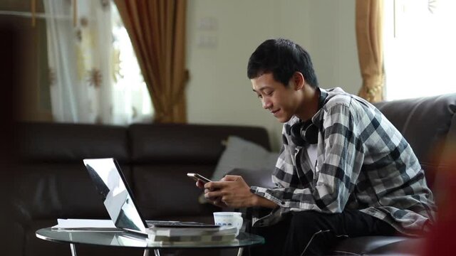 Cheerful of young asian man smiling while chating with mobile phone in front of computer laptop at home