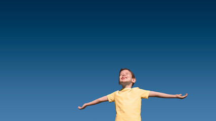 Happy smiling 5-year-old Caucasian boy with closed eyes and upturned face in yellow t-shirt with outstretched arms im summer with blue sky gradient on background. Horizontal banner, large copy space.