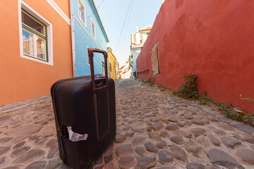 Fototapeta na wymiar Black troler suitcase on an empty medieval city street. Colorful buildings and stone paved street with blue sky. Travel concept.