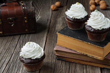 Cupcakes in paper patty cases, topped with whipped cream, are lying on antique books and rustic weathered wood planks. A chest and a few walnut shells are visible in a blurry background.