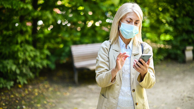 Mature Woman Walking In The Park Using Her Smartphone And Wearing A Mask To Protect From Covid 19 Virus