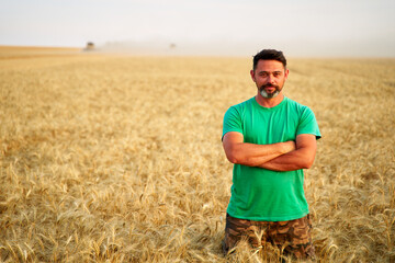 Happy farmer proudly standing in wheat field with arms crossed on chest. Agronomist wearing corporate uniform, looking at camera on farmland. Rich harvest of cultivated cereal crop. Harvesting season.