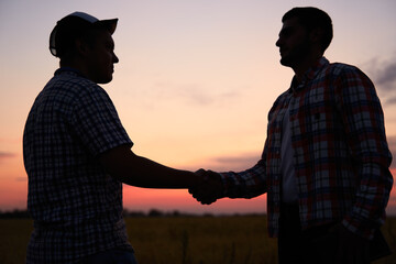 Farmer and agronomist silhouettes shaking hands standing in a wheat field after agreement in dusk....