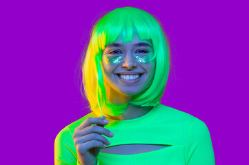 Happy teenage girl dressed in green neon top, wearing yellow funky wig and glitter on cheeks, smiling and laughing at party, isolated on purple background