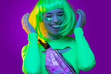 Close-up portrait of young funky girl wearing neon green top, belt bag, yellow wig and glitter on cheeks, laughing and dancing at party, isolated on purple background