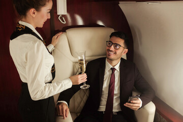 Young billionaire travelling in his private jet holding phone, meeting stewardess who brings him glass of champagne, enjoying business trip