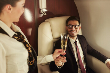 Young business man travelling first class, sitting in suit at table, flight attendant is bringing him glass of champagne to celebrate success