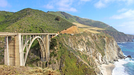 California popular attraction - famous Big Sur coast with the Bixby Creek bridge. The view on the green hills with rocky cliffs and shore with the white sand beach.