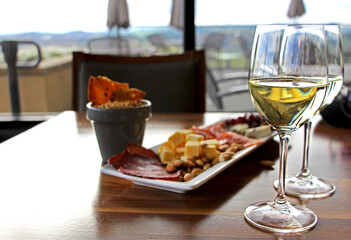Two wine glasses filled with white wine and refreshment plate of selected meat, cheese and nut variation on wooden table - seating area in the background. 