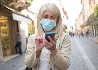 Fototapeta premium Masked woman using her mobile smartphone while walking in a city street during coronavirus pandemic