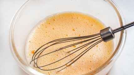 three beaten eggs for cooking French omelet in a glass bowl before whipping. Whisk for whipping lies in a bowl.