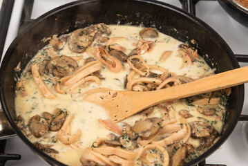 Cooking creamy sauce with squid and mushrooms. Sliced squid rings and champignons, with dill and parsley are stewed with cream in a pan and mixed with a wooden spoon.