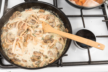 Cooking creamy sauce with squid and mushrooms. Sliced squid rings and champignons, with dill and parsley are stewed with cream in a pan and mixed with a wooden spoon.