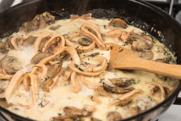 Cooking creamy sauce with squid and mushrooms. Sliced squid rings and champignons, with dill and parsley are stewed with cream in a pan and mixed with a wooden spoon. Close-up