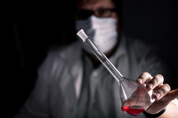 Scientist with white coat and face mask working and testing with red liquid in a conical erlenmeyer flask.