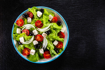 Fresh Greek salad - feta cheese, cherry tomatoes, lettuce, black olives and onion on black stone board
