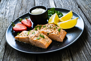 Steamed salmon steaks with lemon, strawberries and creamy dip served on black plate on wooden table
