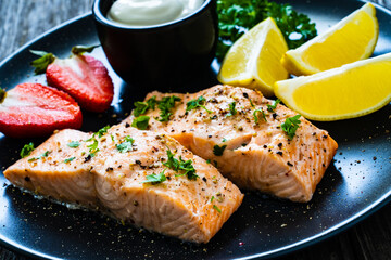 Steamed salmon steaks with lemon, strawberries and creamy dip served on black plate on wooden table
