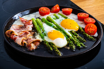 Preparing one pen breakfast. Sunny side up eggs with green asparagus, fried bacon and cherry tomatoes 
