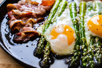 One pen breakfast. Sunny side up eggs with green asparagus, fried bacon and cherry tomatoes on black plate on wooden table

