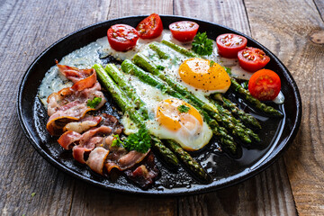 One pen breakfast. Sunny side up eggs with green asparagus, fried bacon and cherry tomatoes on black plate on wooden table
