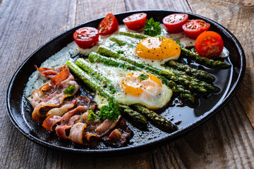 One pen breakfast. Sunny side up eggs with green asparagus, fried bacon and cherry tomatoes on black plate on wooden table
