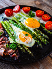 One pen breakfast. Sunny side up eggs with green asparagus, fried bacon and cherry tomatoes on black plate on wooden table
