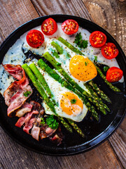 One pen breakfast. Sunny side up eggs with green asparagus, fried bacon and cherry tomatoes on black plate on wooden table
