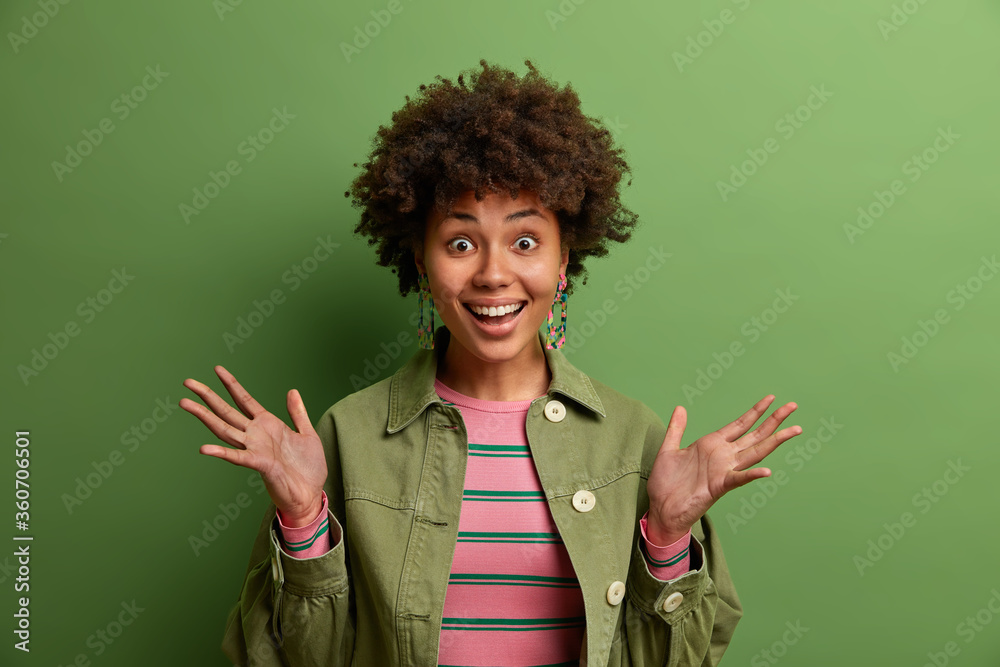 Wall mural Cheerful young woman raises palms and smiles broadly, has happy reaction on something awesome, dressed in fashionable clothes, poses against green background. People, positive emotions, reaction