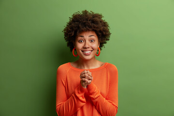 Fototapeta na wymiar Young smiling woman with Afro hair, keeps hands pressed together, looks with friendly pleased look at camera, anticipates something good happen, awaits positive answer from you, models indoor