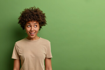 Fototapeta na wymiar Curious cheerful woman with Afro hair looks with interest and happiness aside, has wondered expression, wears casual beige t shirt, isolated on green background, blank copy space on right side