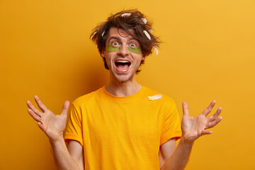 Positive friendly looking man raises hands, happy to meet someone, awakes in morning to have skin facial treatment, applies collagen patches under eyes, dressed in yellow t shirt. Good morning concept