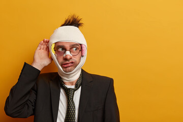 Pensive young man keeps hand on hurt head, looks with worried expression aside, thinks how to...