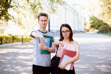 Two angry and bewildered friends holding learning materials gesturing thumbs down in the street...