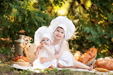 Healthy eating concept. Happy little girl and boy cook outdoors on a sunny summer day. Toddler baker on a picnic eats bread and bagels in a white apron and hat