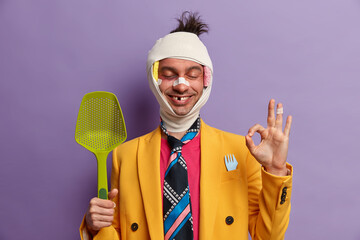Studio shot of happy positive man with various traumas after ridiculous accident, makes okay...