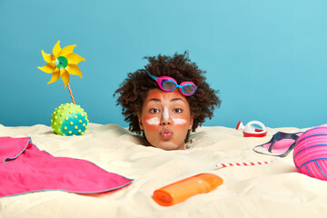 Outdoor shot of young curly woman in romantic mood, spends time at beach, applies sunscreen cream...