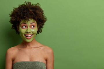 Lovely happy woman with curly hair, looks aside smiling, wears green beauty mask for skin pampering, cares about complexion, stands wrapped in bath towel, bare shoulders, isolated on green background