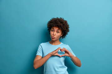 Romantic beautiful curly haired young woman expresses feelings with amorous heart gesture,...