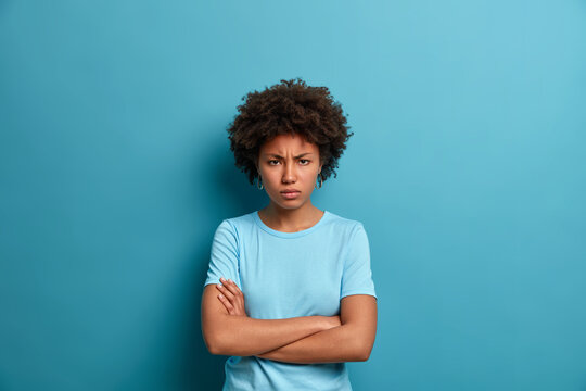Horizontal shot of resentful offended woman crosses hands over body, looks at camera with angry gaze and sullen expression, being angry with someone, has formidable look, poses over blue studio wall